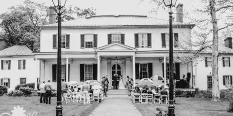 Black and white photo of a wedding ceremony taking place on the front lawn of a historic white Georgian mansion with a portico.