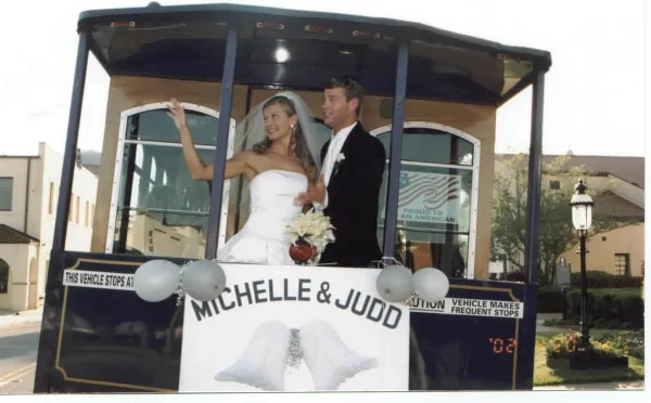 A bride and groom standing on the rear platform of a trolley decorated with balloons and a sign reading 'Michelle & Judd'.
