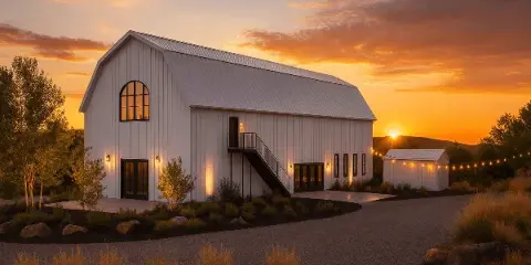 Exterior view of a white barn-style wedding venue at sunset with a gravel driveway and string lights.