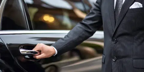 A professional chauffeur in a dark suit opening the door of a black luxury car.