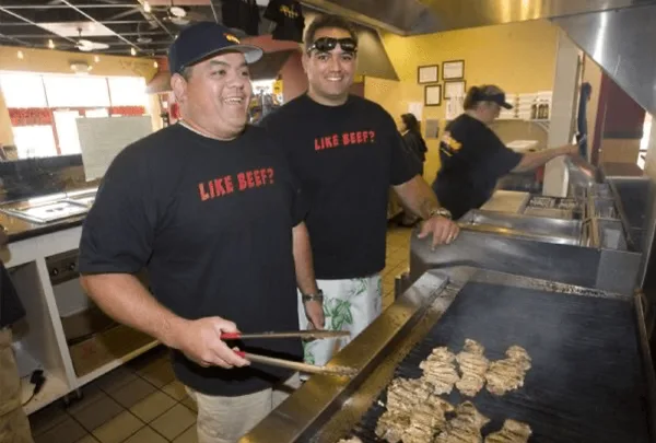 Two restaurant employees wearing black t-shirts stand behind a flat-top grill cooking food in a commercial kitchen.