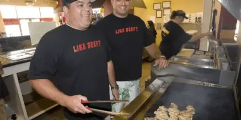 Two restaurant employees wearing black t-shirts stand behind a flat-top grill cooking food in a commercial kitchen.