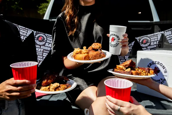 A person holding a plate of fried chicken and sausage next to a white cup with the Bergeron's Boudin & Cajun Meats logo, with branded bags and other food plates visible in the background.