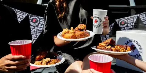 A person holding a plate of fried chicken and sausage next to a white cup with the Bergeron's Boudin & Cajun Meats logo, with branded bags and other food plates visible in the background.