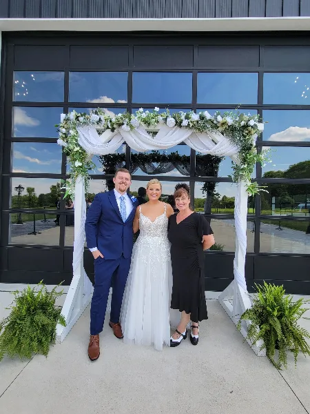 A bride and groom posing with a woman in a black dress under a floral wedding arch.
