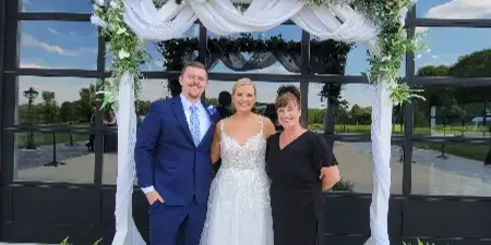 A bride and groom posing with a woman in a black dress under a floral wedding arch.