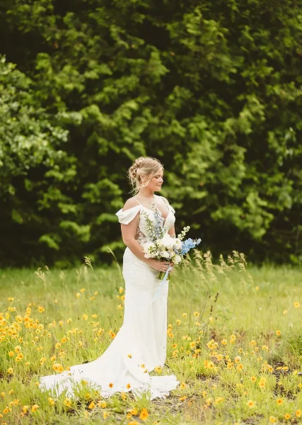 A bride in a white wedding dress holding a bouquet of white and blue flowers in a grassy field.