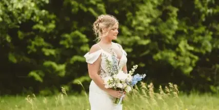 A bride in a white wedding dress holding a bouquet of white and blue flowers in a grassy field.