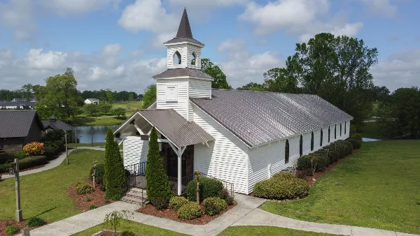 A white wooden chapel with a steeple and metal roof, surrounded by green lawns and trees, with a pond visible in the background.