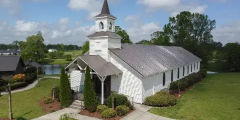 A white wooden chapel with a steeple and metal roof, surrounded by green lawns and trees, with a pond visible in the background.