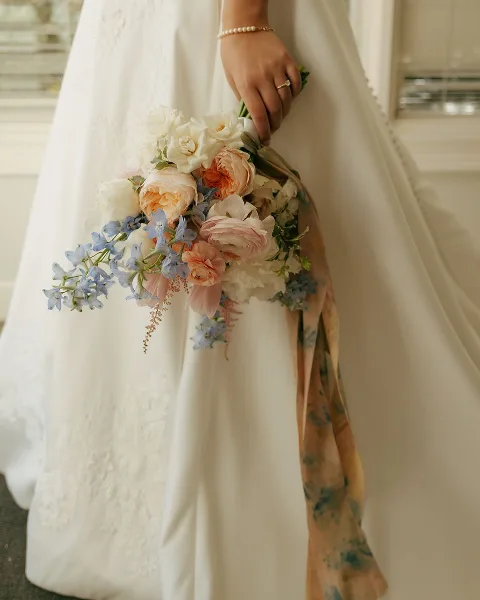 A close-up of a bride's hand holding a cascading bouquet of white and peach roses, ranunculus, and blue delphiniums against a white wedding dress.