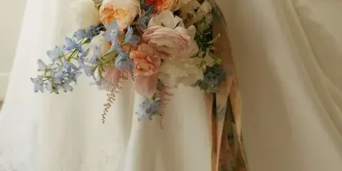 A close-up of a bride's hand holding a cascading bouquet of white and peach roses, ranunculus, and blue delphiniums against a white wedding dress.