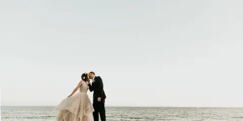 A bride and groom kissing on a stone pier overlooking the ocean.
