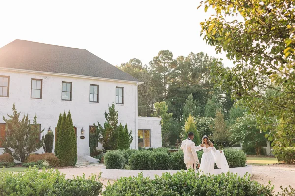 A bride and groom standing in a garden in front of a white brick building during a wedding.