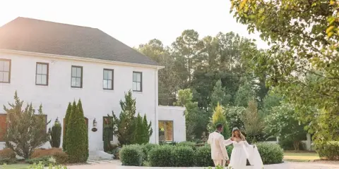A bride and groom standing in a garden in front of a white brick building during a wedding.