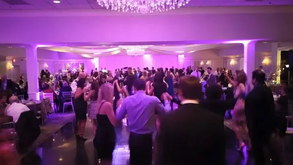 A crowded wedding dance floor with guests dancing under purple lighting and chandeliers.