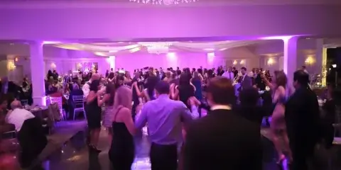 A crowded wedding dance floor with guests dancing under purple lighting and chandeliers.