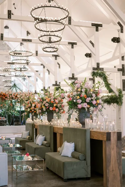 A wedding reception venue featuring a lounge area with green velvet sofas, a wooden bar, and large floral centerpieces under a high ceiling with circular chandeliers.