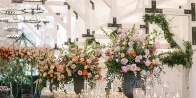 A wedding reception venue featuring a lounge area with green velvet sofas, a wooden bar, and large floral centerpieces under a high ceiling with circular chandeliers.