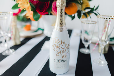 A styled event table featuring a custom-labeled champagne bottle, floral centerpiece, and gold-rimmed glassware on a black and white striped tablecloth.