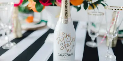 A styled event table featuring a custom-labeled champagne bottle, floral centerpiece, and gold-rimmed glassware on a black and white striped tablecloth.