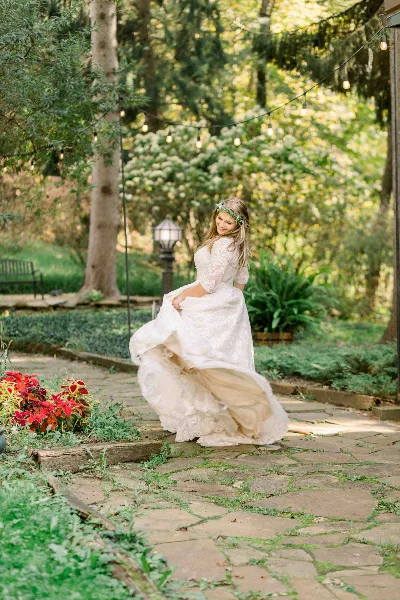 A bride in a white lace wedding dress and floral crown twirling on a stone path in a garden setting.