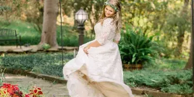 A bride in a white lace wedding dress and floral crown twirling on a stone path in a garden setting.