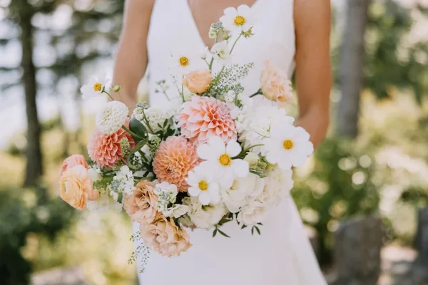 A bride in a white dress holding a large bouquet of peach, white, and yellow flowers.
