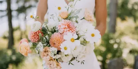 A bride in a white dress holding a large bouquet of peach, white, and yellow flowers.