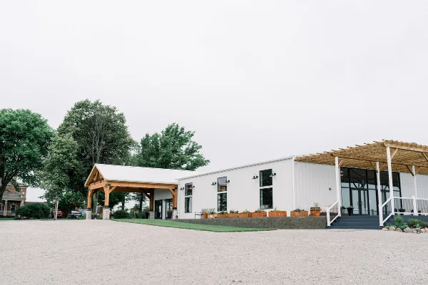 Exterior view of The 1868 Farmhouse wedding venue, showing a white metal building with a wooden pergola and covered entryway.