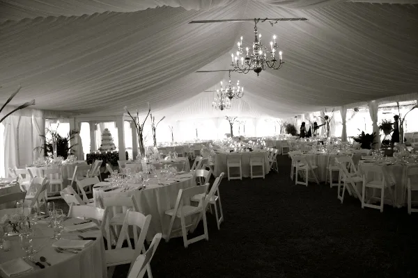 Interior of a white wedding tent with draped ceiling, hanging chandeliers, round tables with white linens, and white chairs arranged for a reception.