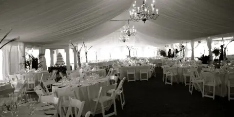 Interior of a white wedding tent with draped ceiling, hanging chandeliers, round tables with white linens, and white chairs arranged for a reception.