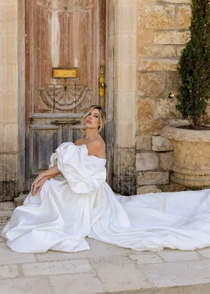 A bride with styled hair and makeup sitting in a white wedding dress in front of a rustic stone wall and wooden door.
