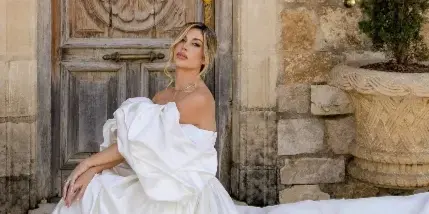 A bride with styled hair and makeup sitting in a white wedding dress in front of a rustic stone wall and wooden door.