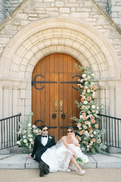 A bride and groom sitting on steps in front of a stone archway decorated with a large floral arrangement of peach and white roses and greenery.