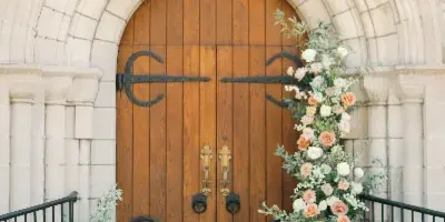 A bride and groom sitting on steps in front of a stone archway decorated with a large floral arrangement of peach and white roses and greenery.