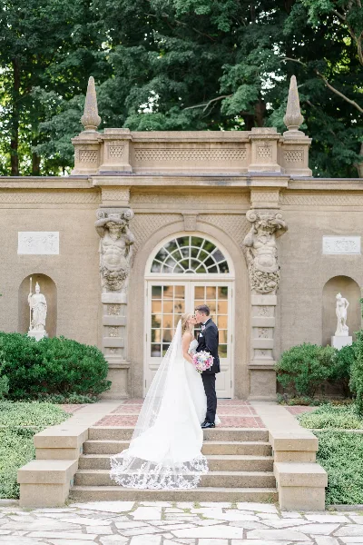 A bride and groom kissing in front of an ornate stone building entrance with classical statues and manicured landscaping.