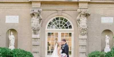 A bride and groom kissing in front of an ornate stone building entrance with classical statues and manicured landscaping.