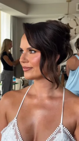A woman with a professional updo hairstyle and makeup, wearing a white lace top, in a room with other people in the background.
