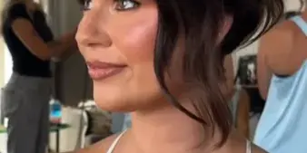 A woman with a professional updo hairstyle and makeup, wearing a white lace top, in a room with other people in the background.