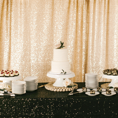 A wedding dessert table with a white tiered cake, stacked white plates, and a gold sequin backdrop.