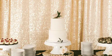 A wedding dessert table with a white tiered cake, stacked white plates, and a gold sequin backdrop.