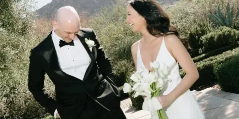 A bride holding a white bouquet and a groom wearing a boutonniere standing outdoors.