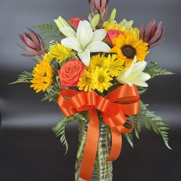 A mixed floral bouquet featuring white lilies, yellow daisies, sunflowers, and a red rose, arranged in a clear glass vase with an orange ribbon.