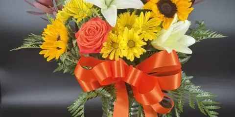 A mixed floral bouquet featuring white lilies, yellow daisies, sunflowers, and a red rose, arranged in a clear glass vase with an orange ribbon.