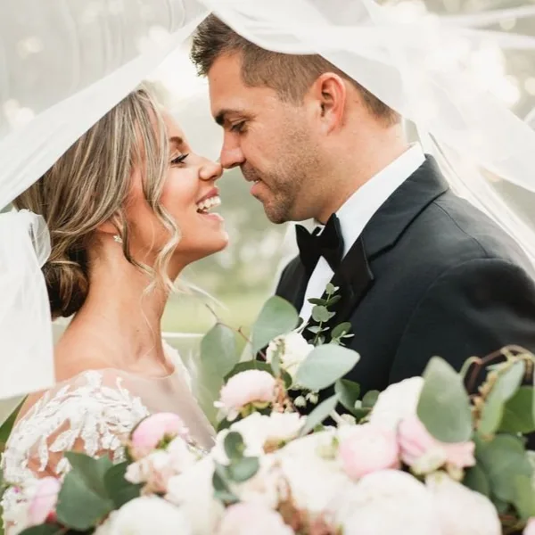 A bride and groom smiling at each other under a veil, with a large bouquet of white and pink flowers and greenery in the foreground.