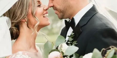 A bride and groom smiling at each other under a veil, with a large bouquet of white and pink flowers and greenery in the foreground.