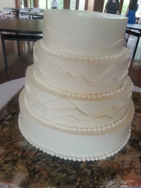 A four-tier white wedding cake with piped bead borders and wavy fondant accents, displayed on a granite countertop.