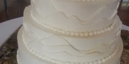 A four-tier white wedding cake with piped bead borders and wavy fondant accents, displayed on a granite countertop.