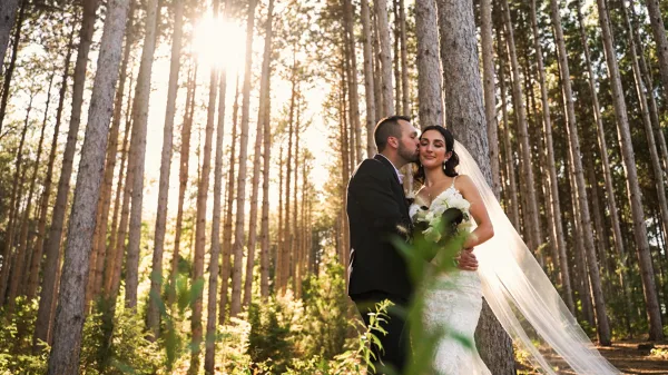 A bride and groom embracing in a sunlit forest, with the bride holding a bouquet and wearing a veil.